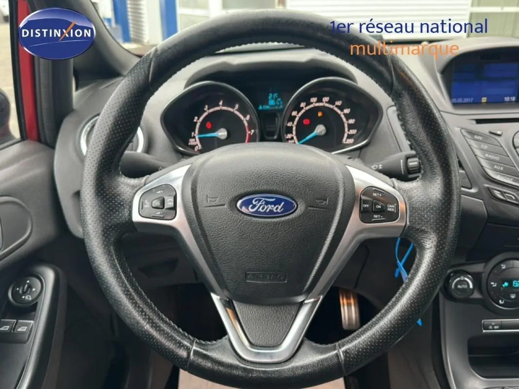 Vue centrée sur le volant noir perforé de la Ford Fiesta rouge, avec tableau de bord et commandes visibles.