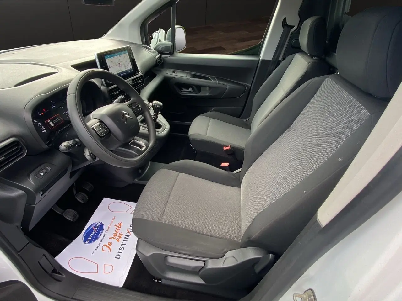 Vue intérieure côté conducteur du Citroën Berlingo blanc, montrant le tableau de bord et les sièges en tissu gris.