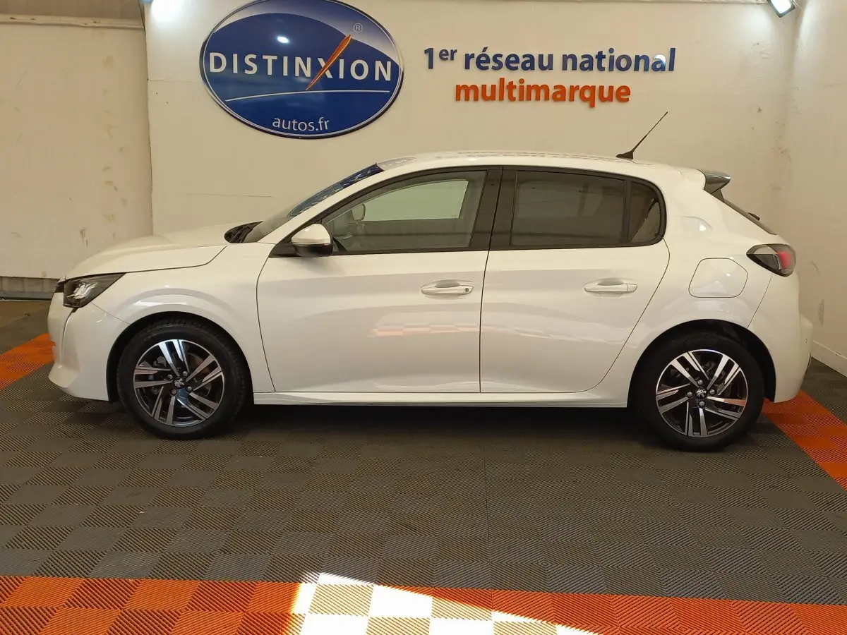 Profil côté gauche d'une Peugeot 208 PureTech 100 S&S Allure blanche, en intérieur sur sol gris et orange.