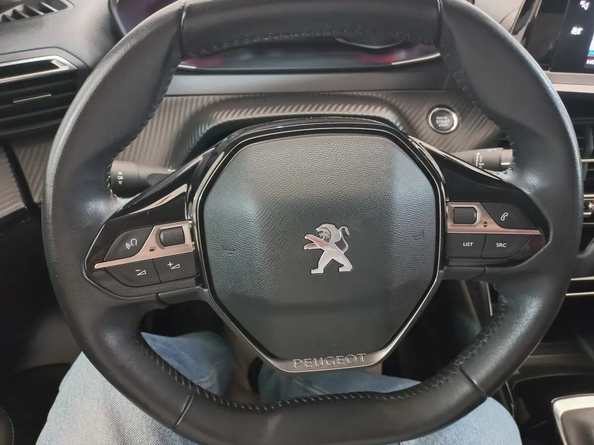 Vue rapprochée du volant cuir noir de la Peugeot 208 PureTech 100 S&S Allure avec commandes intégrées et bouton démarrage visible.