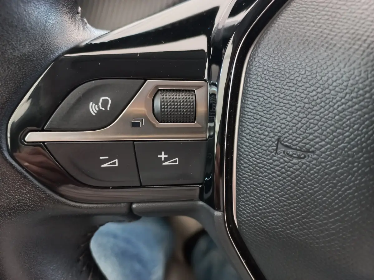 Gros plan sur les commandes de volume et reconnaissance vocale sur le volant cuir noir d'une Peugeot 208 blanche.