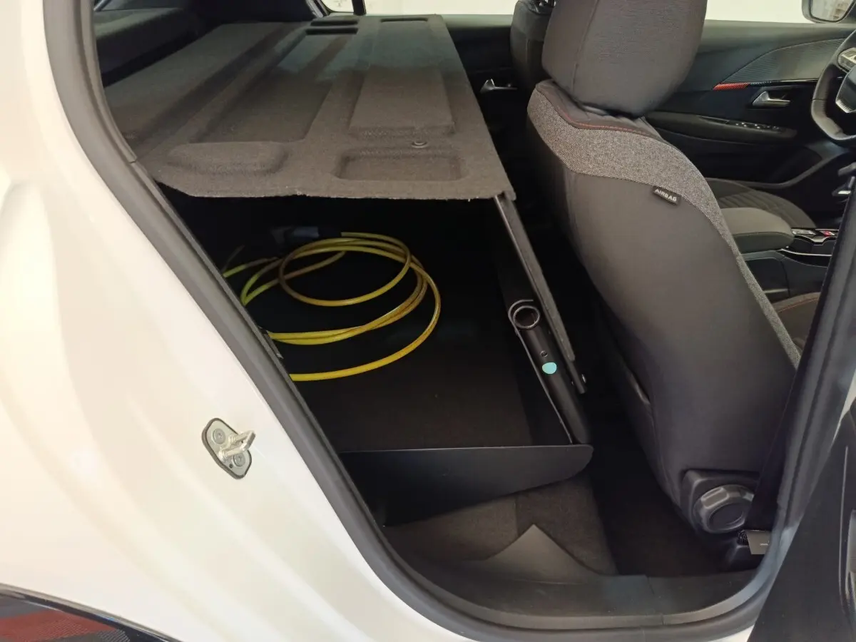 Vue intérieure côté arrière droit d'une Peugeot 208 électrique blanche, montrant le rangement sous la banquette avec câble de charge.