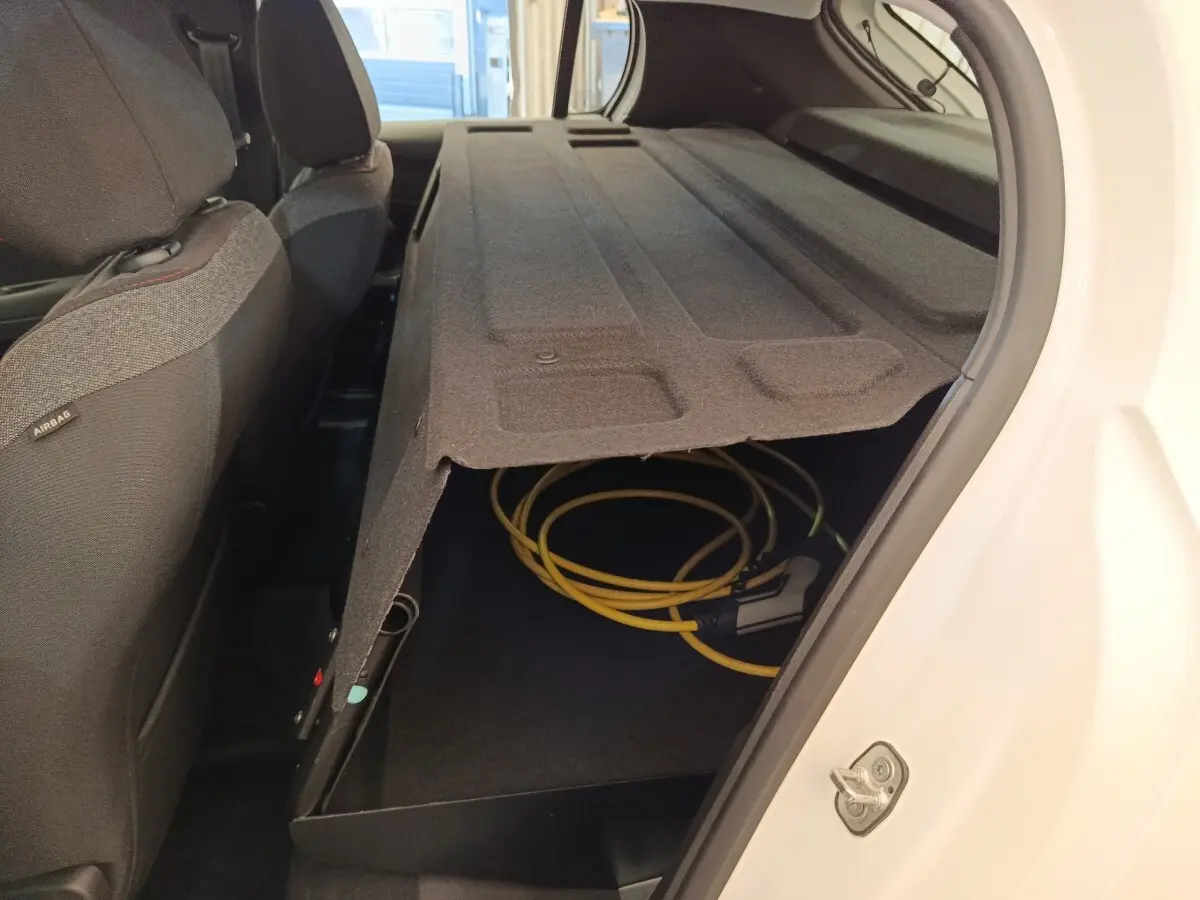 Vue intérieure arrière droite du coffre d'une Peugeot 208 électrique blanche avec câble de recharge rangé sous le plancher.