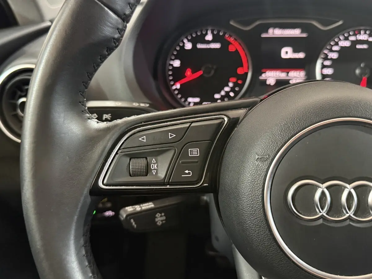 Gros plan sur le volant cuir noir d'Audi A3 2.0 TDI 2018 avec commandes multifonctions et compteurs en arrière-plan flous.