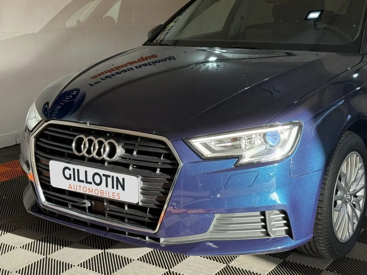 Vue 3/4 avant droit d'une Audi A3 bleu métallisé avec calandre hexagonale et phares LED allumés.