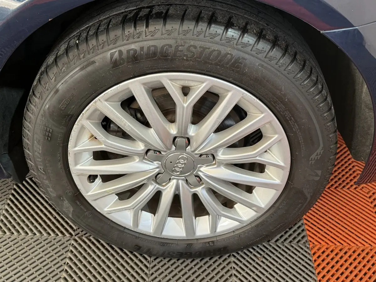 Gros plan sur la jante alliage argentée avec logo Audi d'une Audi A3 bleu, pneu Bridgestone visible au-dessus d'un sol carrelé.