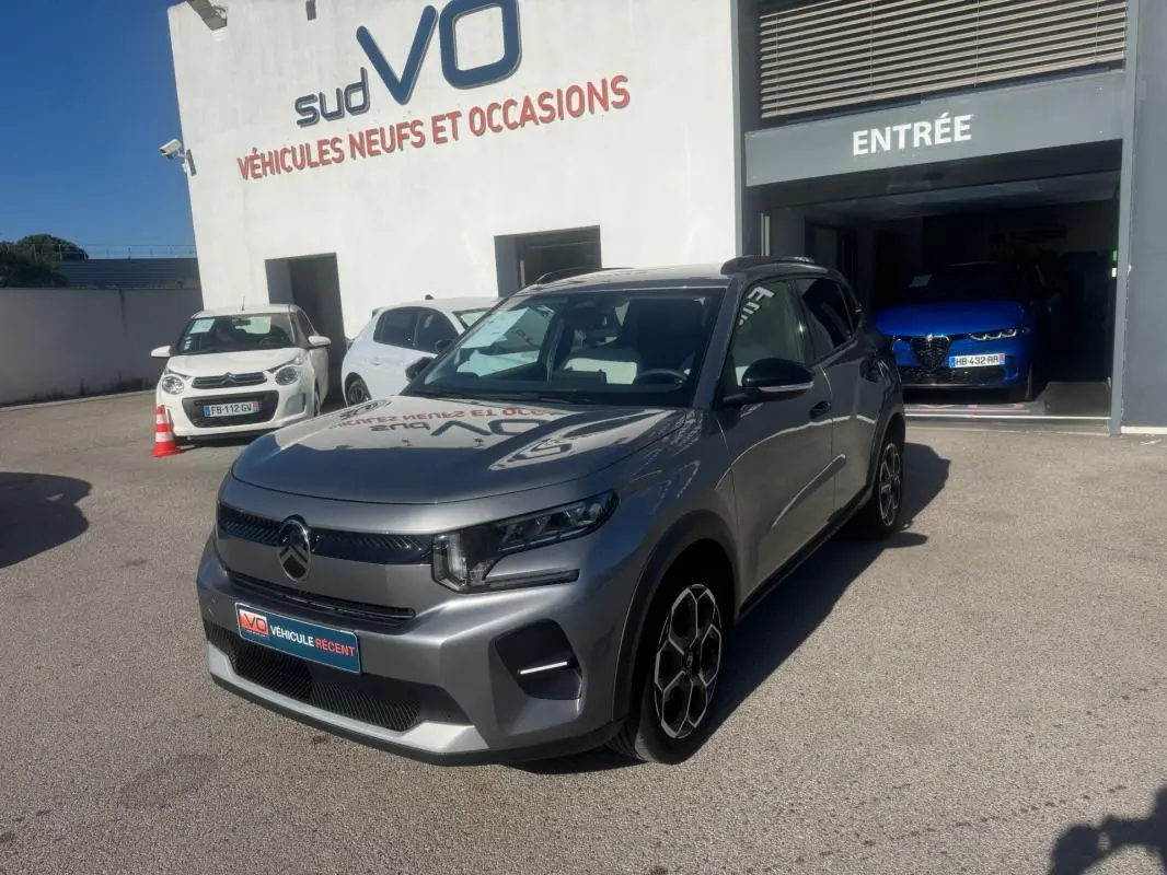 Citroën C3 gris 3/4 avant droit avec jantes alliage et barres de toit, stationnée devant un garage.