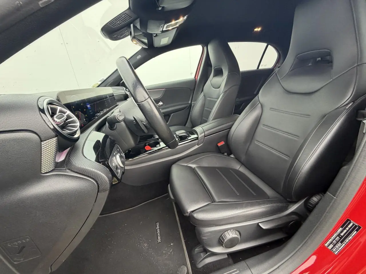 Vue intérieure côté conducteur de la Mercedes Classe A rouge, sièges cuir noir et tableau de bord moderne avec écran tactile.
