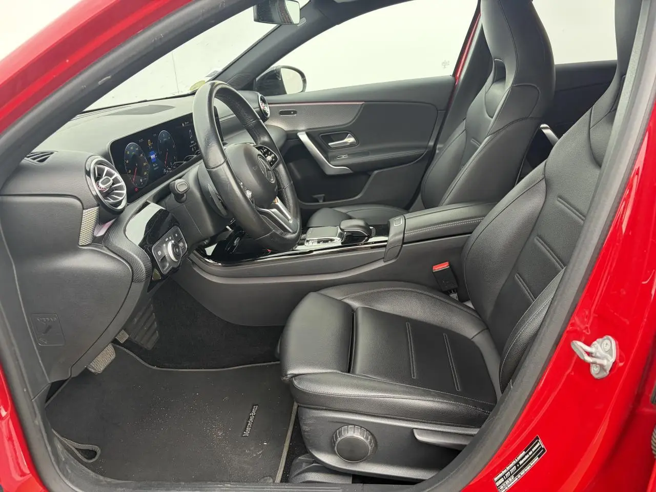 Vue intérieure côté conducteur d'une Mercedes Classe A rouge, mettant en valeur les sièges cuir noirs et le tableau de bord moderne.