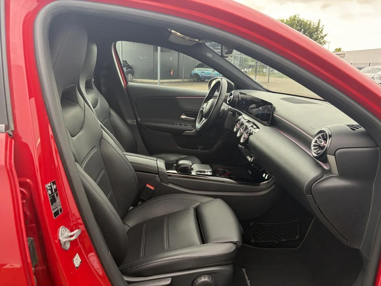 Vue intérieure côté conducteur d'une Mercedes Classe A rouge, avec sellerie cuir noir et tableau de bord moderne.