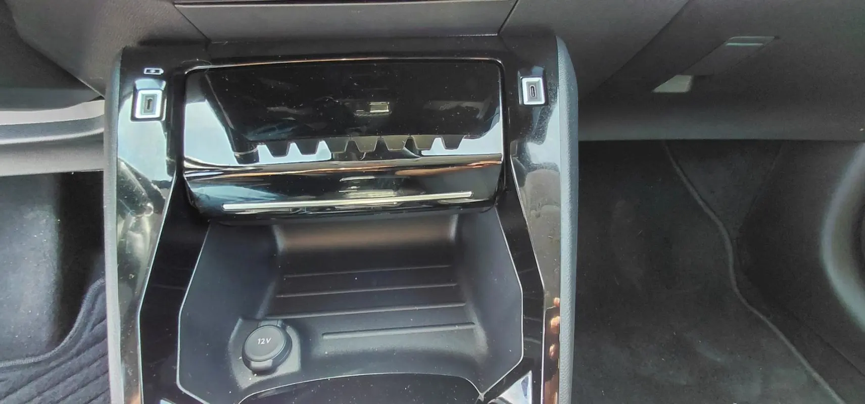 Gros plan sur la console centrale noire brillante de la Peugeot 208 Hybrid 110 ch, avec prise 12V visible.