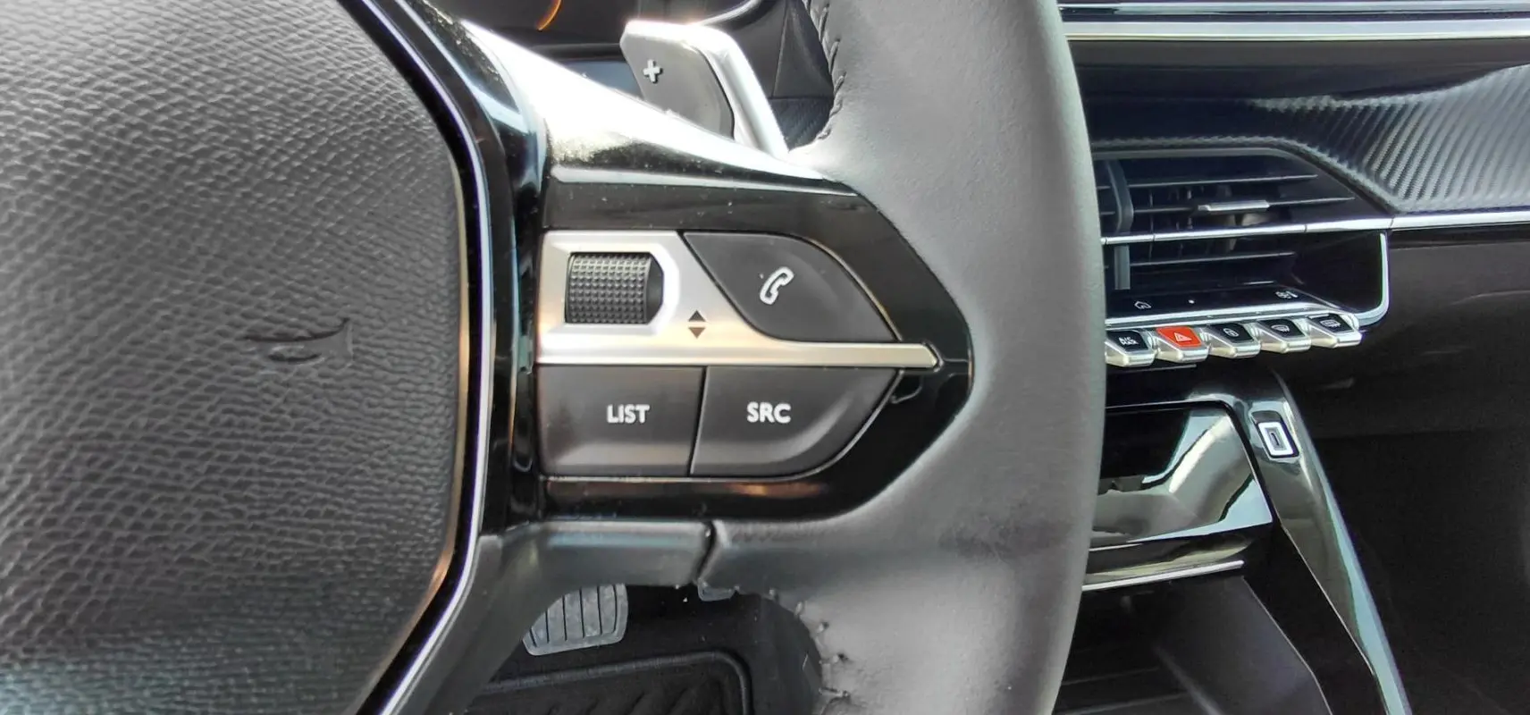 Gros plan sur les commandes gauche du volant cuir noir de la Peugeot 208 Hybrid 110 ch, avec tableau de bord et aérateurs en arrière-plan.