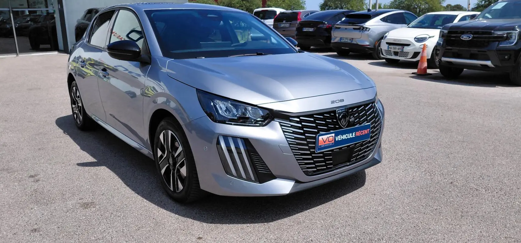 Peugeot 208 gris vue 3/4 avant droit, avec calandre noire distinctive et jantes alliage 16 pouces.
