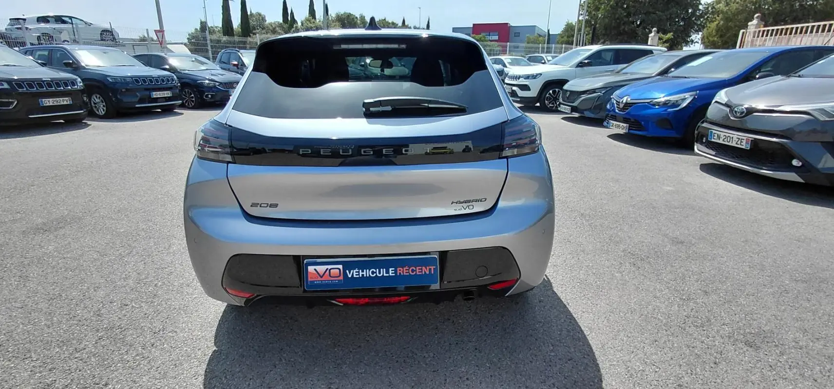 Vue arrière d'une Peugeot 208 gris clair Hybrid 110 ch avec feux noirs et plaque "Véhicule récent" sur parking.