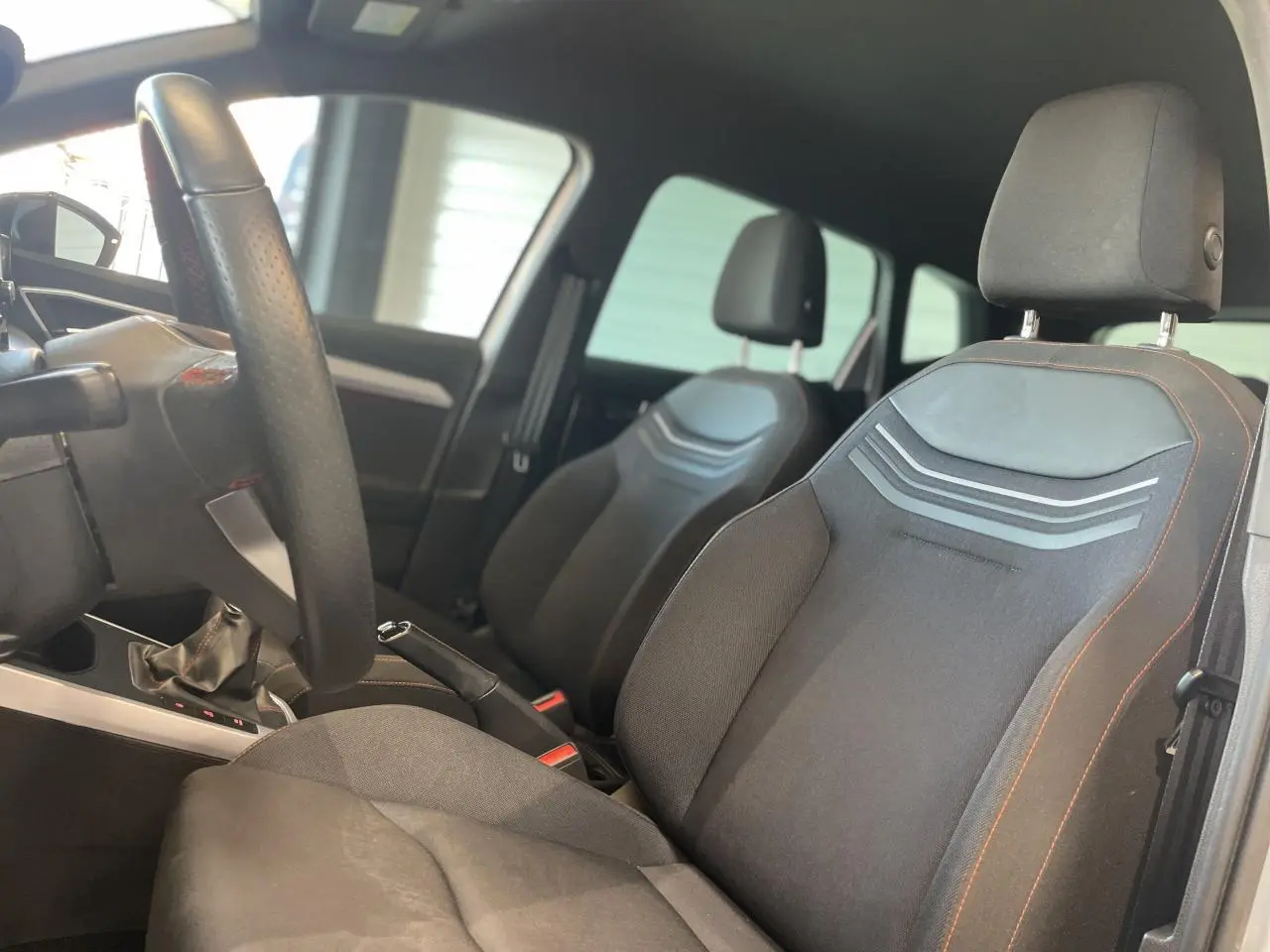 Vue intérieure côté conducteur du SEAT Arona 2024, sièges textile noirs avec surpiqûres orange et volant sport cuir.