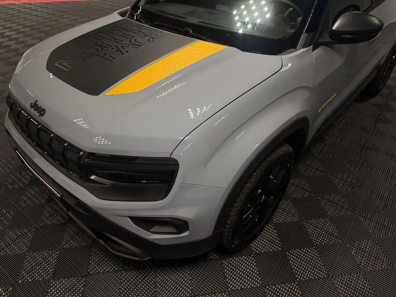 Vue 3/4 avant droit d’un Jeep Avenger gris avec stickers The North Face noirs et jaunes sur le capot et jantes noires.