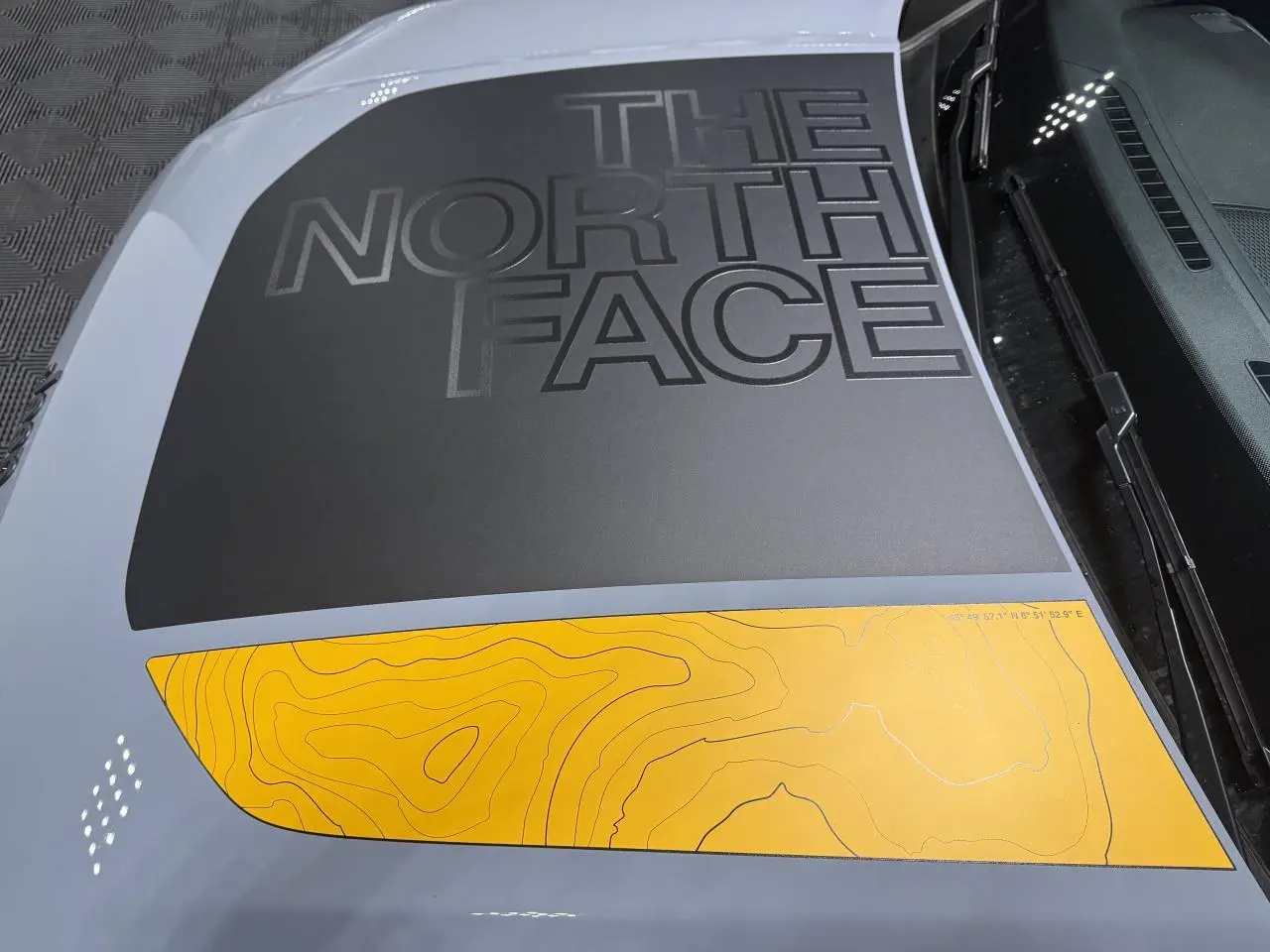 Vue rapprochée du capot gris du Jeep Avenger avec sticker noir "The North Face" et motif topographique jaune.