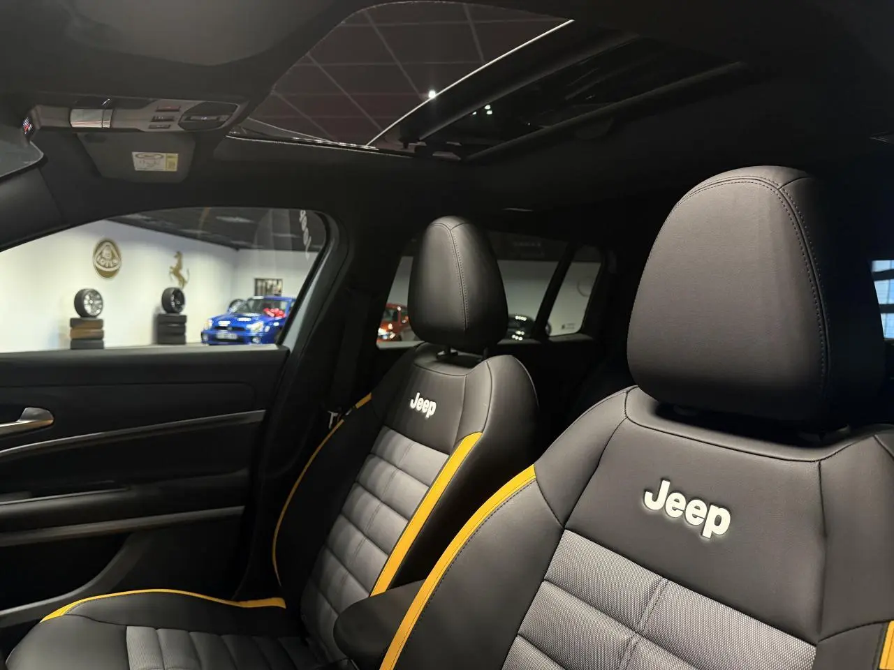 Vue intérieure des sièges avant noirs et gris avec surpiqûres jaunes et logo Jeep, toit ouvrant en verre visible au-dessus.