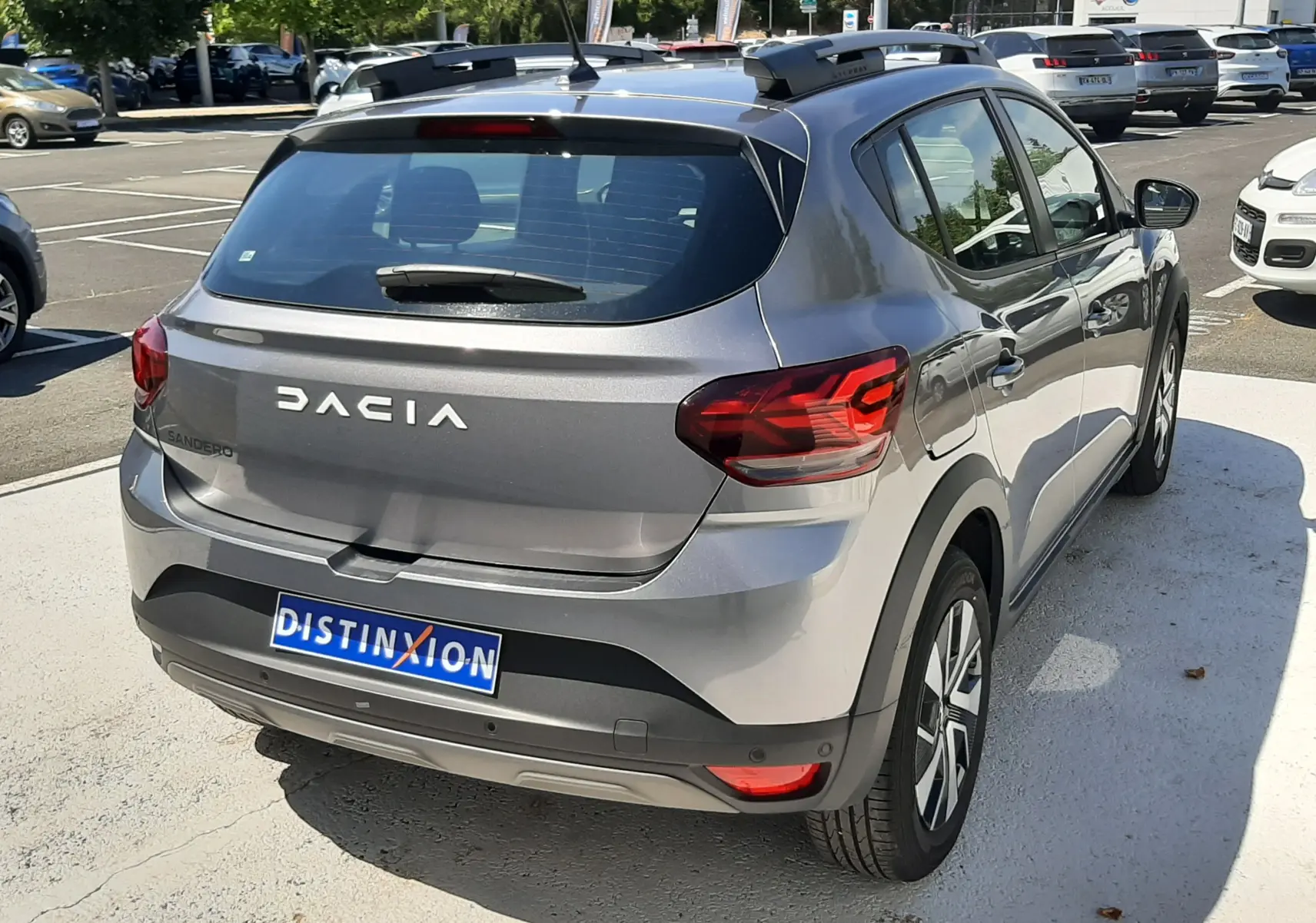 Vue 3/4 arrière droite d'une Dacia Sandero Stepway gris schiste avec barres de toit et feux arrière LED sur parking.