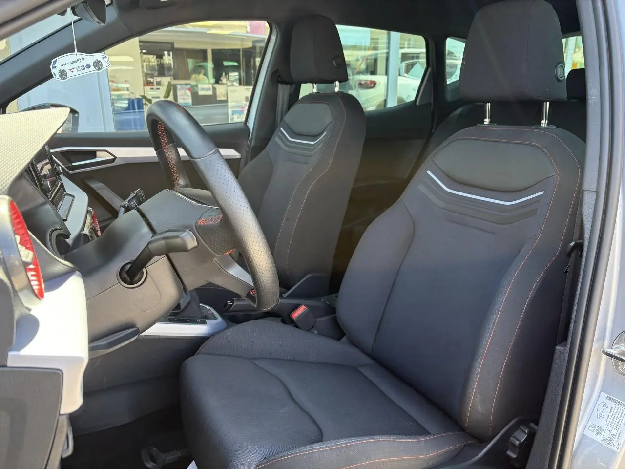 Vue intérieure côté conducteur du SEAT Arona 2024 avec sièges textile noirs et volant sport cuir à surpiqûres rouges.