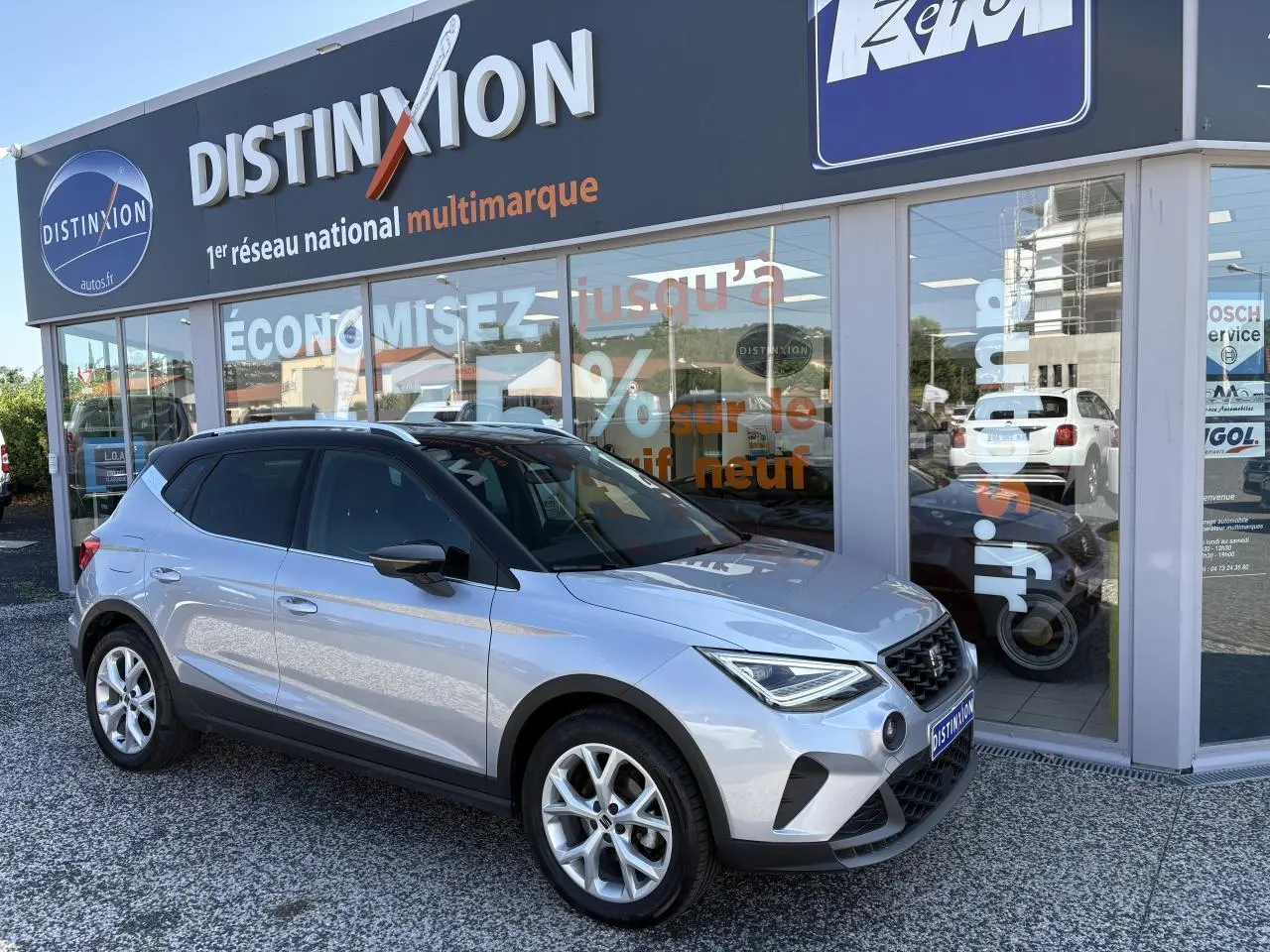 SEAT Arona 2024 gris clair en 3/4 avant droit, avec jantes alliage et toit noir devant concession multimarque.