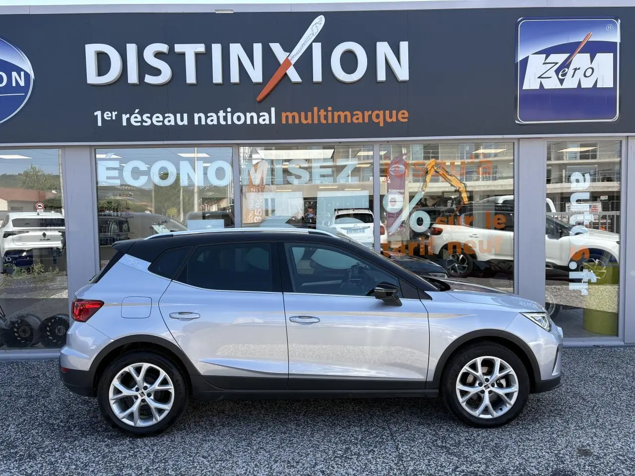 Vue de profil droit d’un SEAT Arona 2024 gris clair avec toit noir et jantes alliage 16 pouces devant un showroom Distinxion.