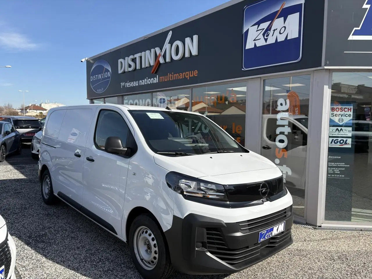 Opel Vivaro blanc en 3/4 avant droit, avec calandre noire et pare-chocs avant noir mat, stationné devant un showroom.