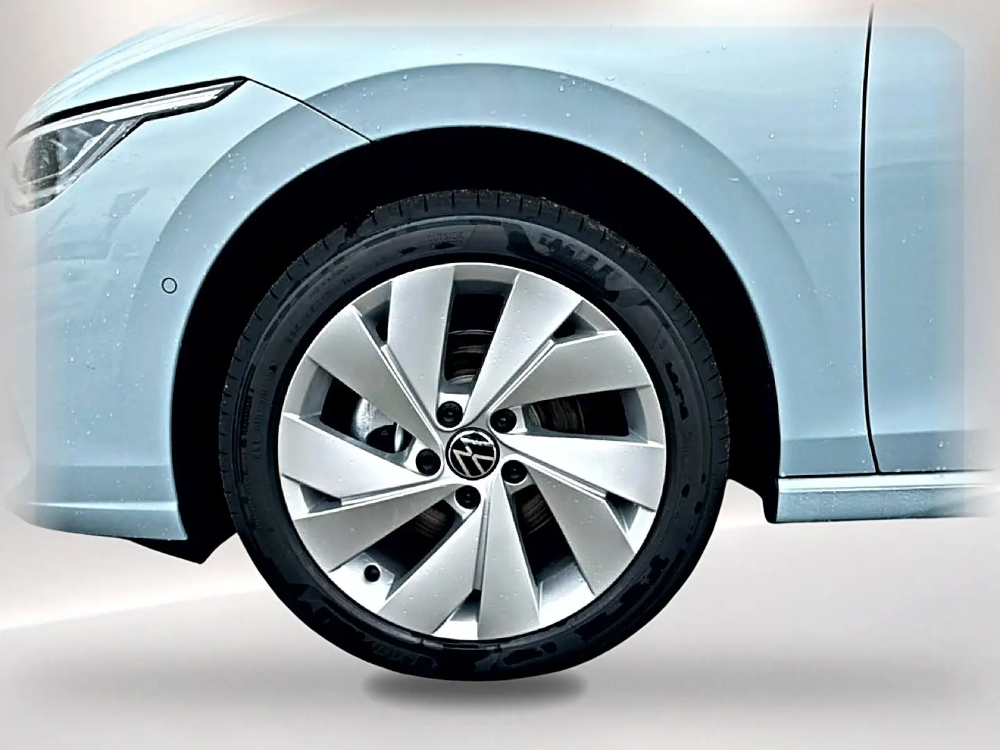 Gros plan sur la roue avant gauche d’une Volkswagen Golf bleu glacier avec jante alu 17 pouces et phare avant partiel visible.