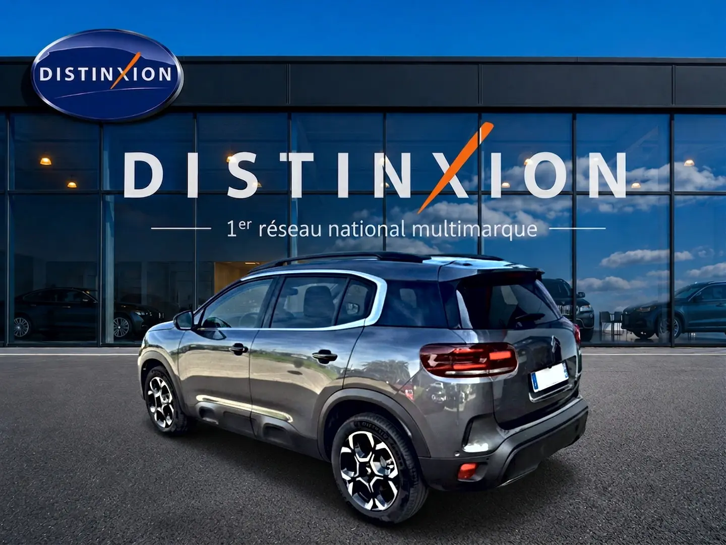 Citroën C5 Aircross gris vue 3/4 arrière droit, avec jantes alliage et barres de toit visibles devant un showroom Distinxion.