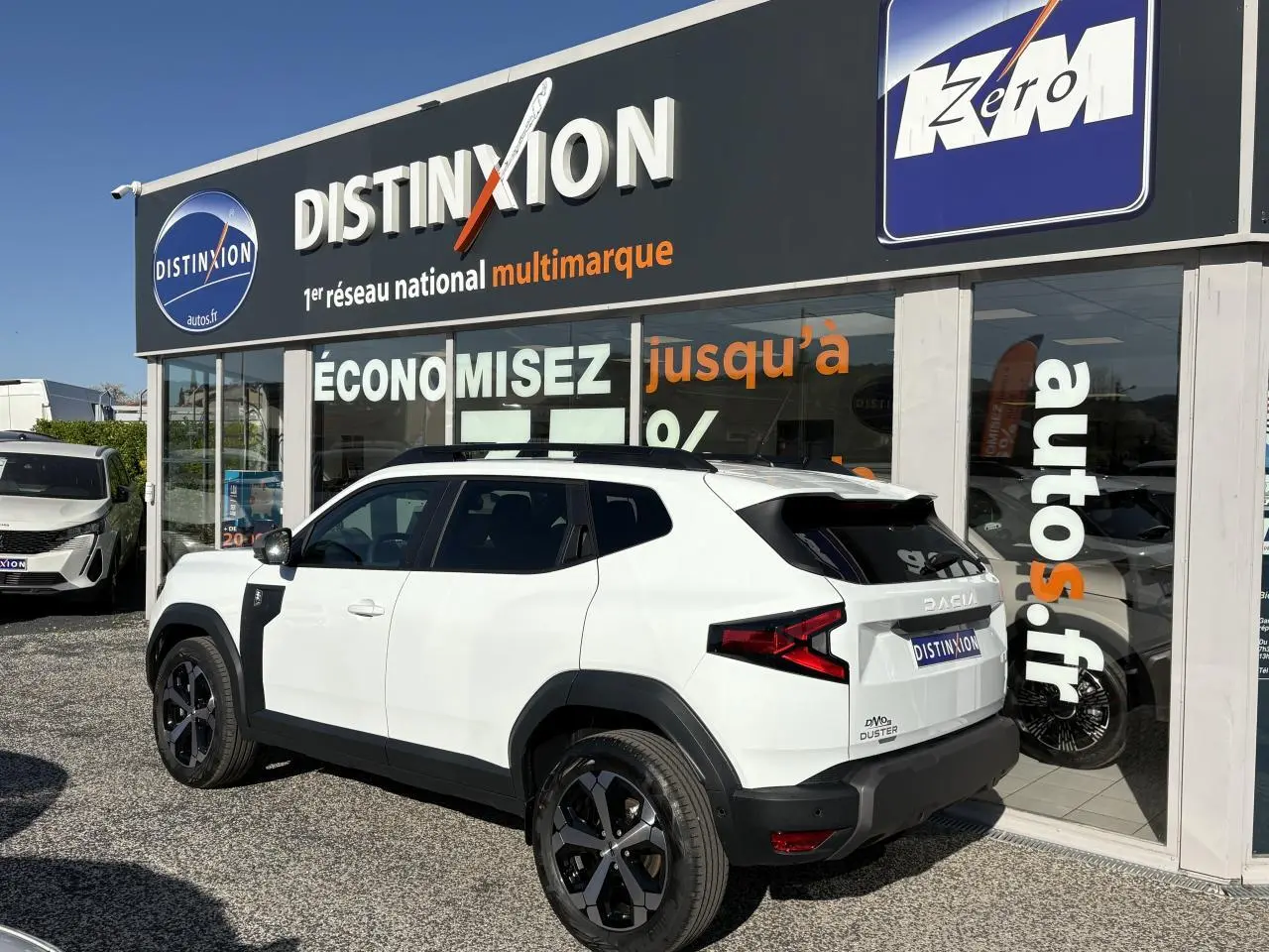 Vue 3/4 arrière droite d'un Dacia Duster blanc métallisé avec barres de toit noires devant une concession Distinxion.