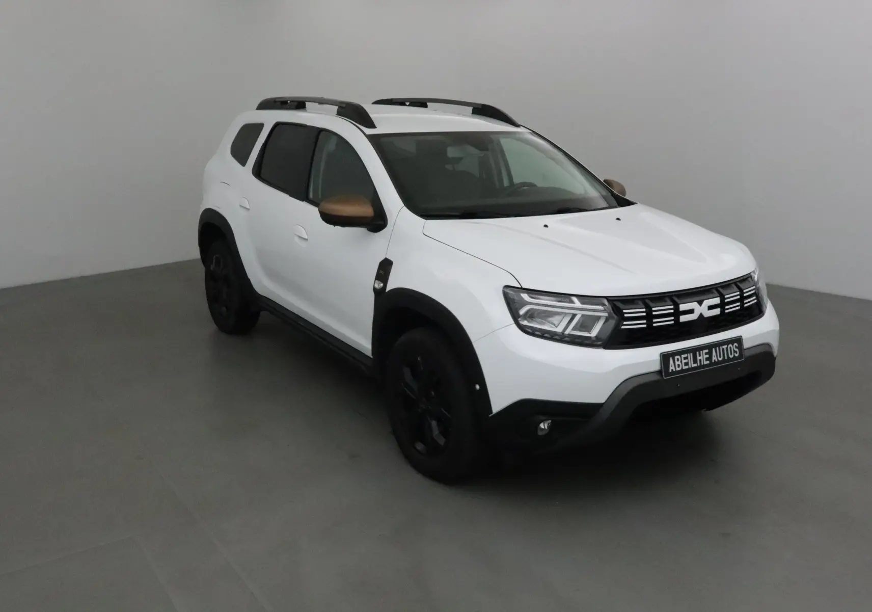 Dacia Duster 4x4 blanc en 3/4 avant droit avec rétroviseurs bronze et jantes noires dans un studio gris.