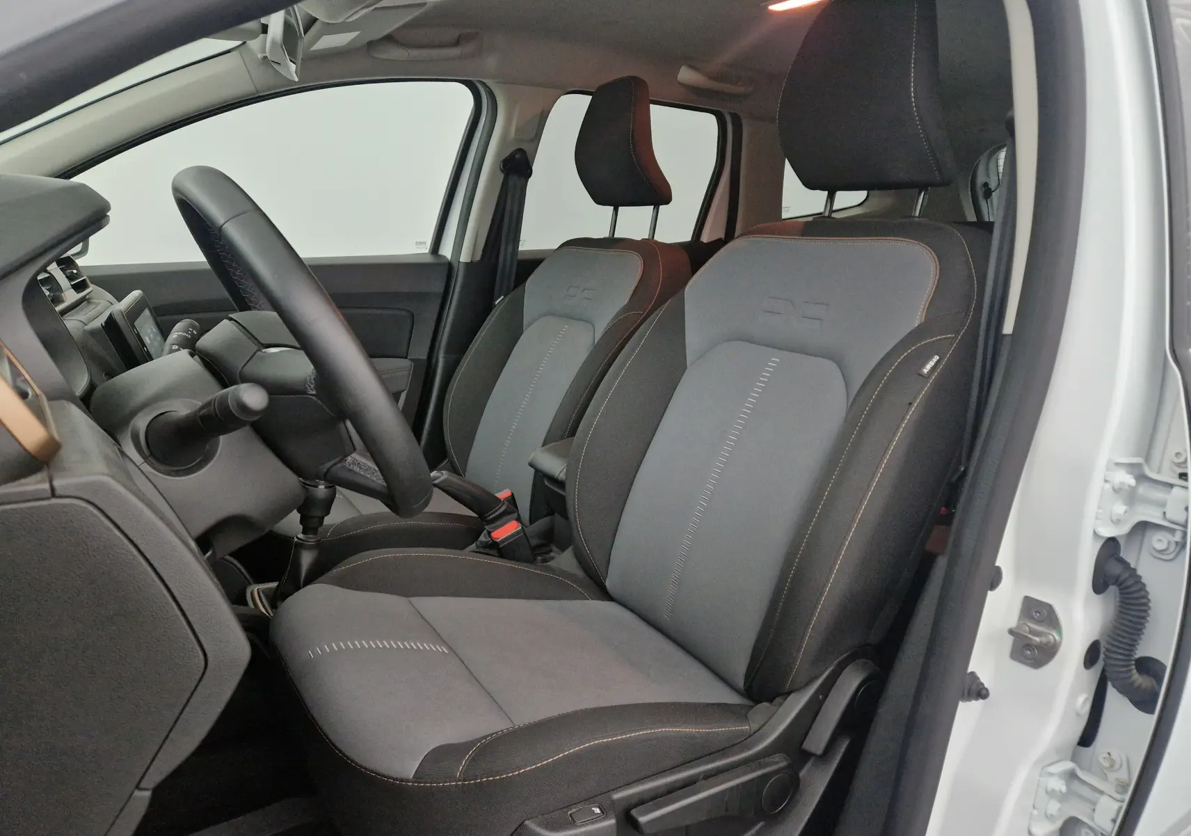 Vue intérieure côté conducteur du Dacia Duster blanc 2024, mettant en valeur les sièges bi-matière gris et noirs avec surpiqûres contrastées.