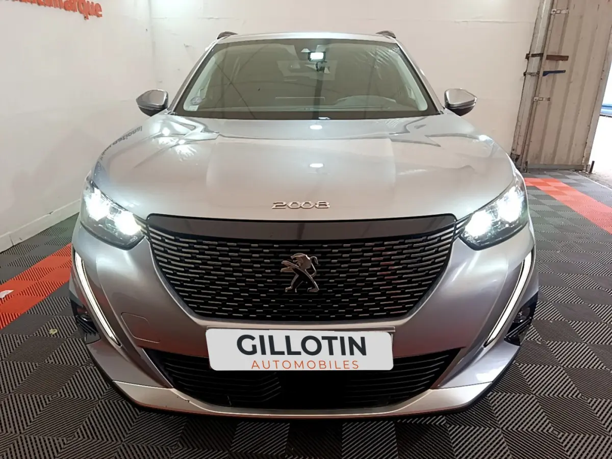 Vue frontale d'un Peugeot 2008 gris clair avec phares allumés et calandre noire distinctive dans un garage.
