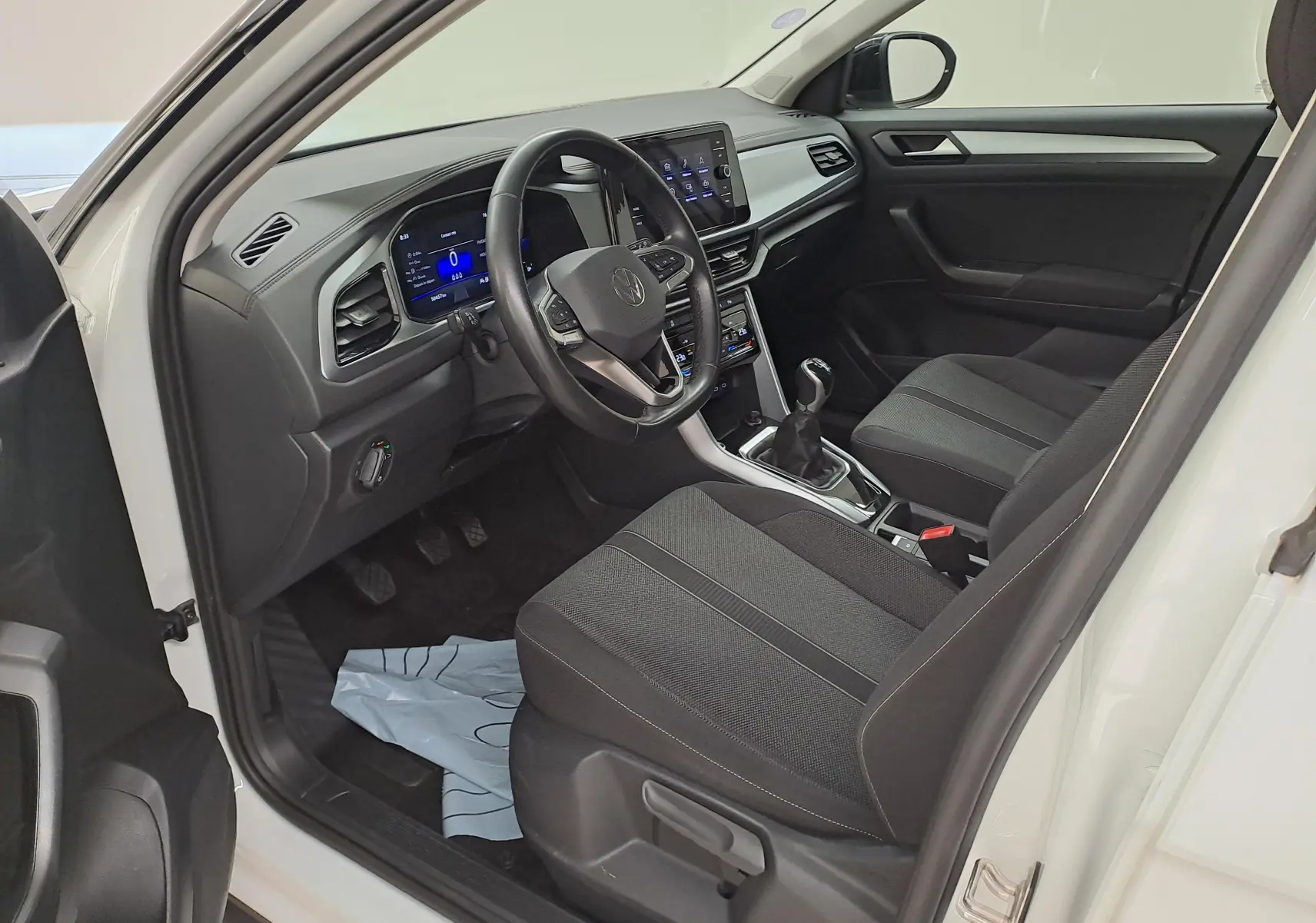 Vue intérieure côté conducteur du Volkswagen T-Roc blanc toit noir, montrant le tableau de bord numérique et la boîte manuelle.