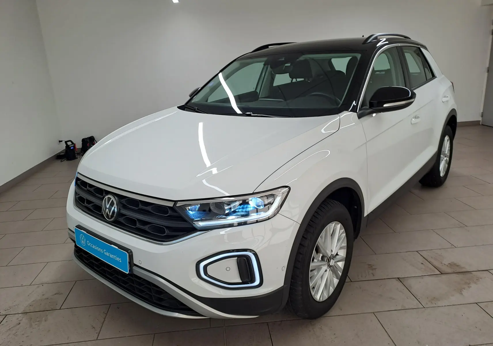 Volkswagen T-Roc blanc pur avec toit noir, vue 3/4 avant droit, phares allumés dans un intérieur lumineux.