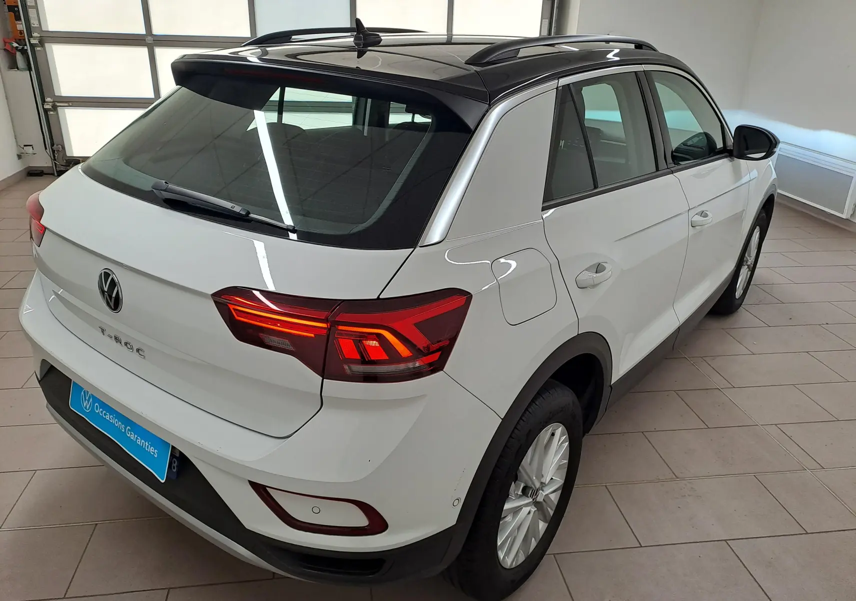 Vue 3/4 arrière droite du Volkswagen T-Roc blanc pur avec toit noir, feux arrière allumés, en intérieur showroom.