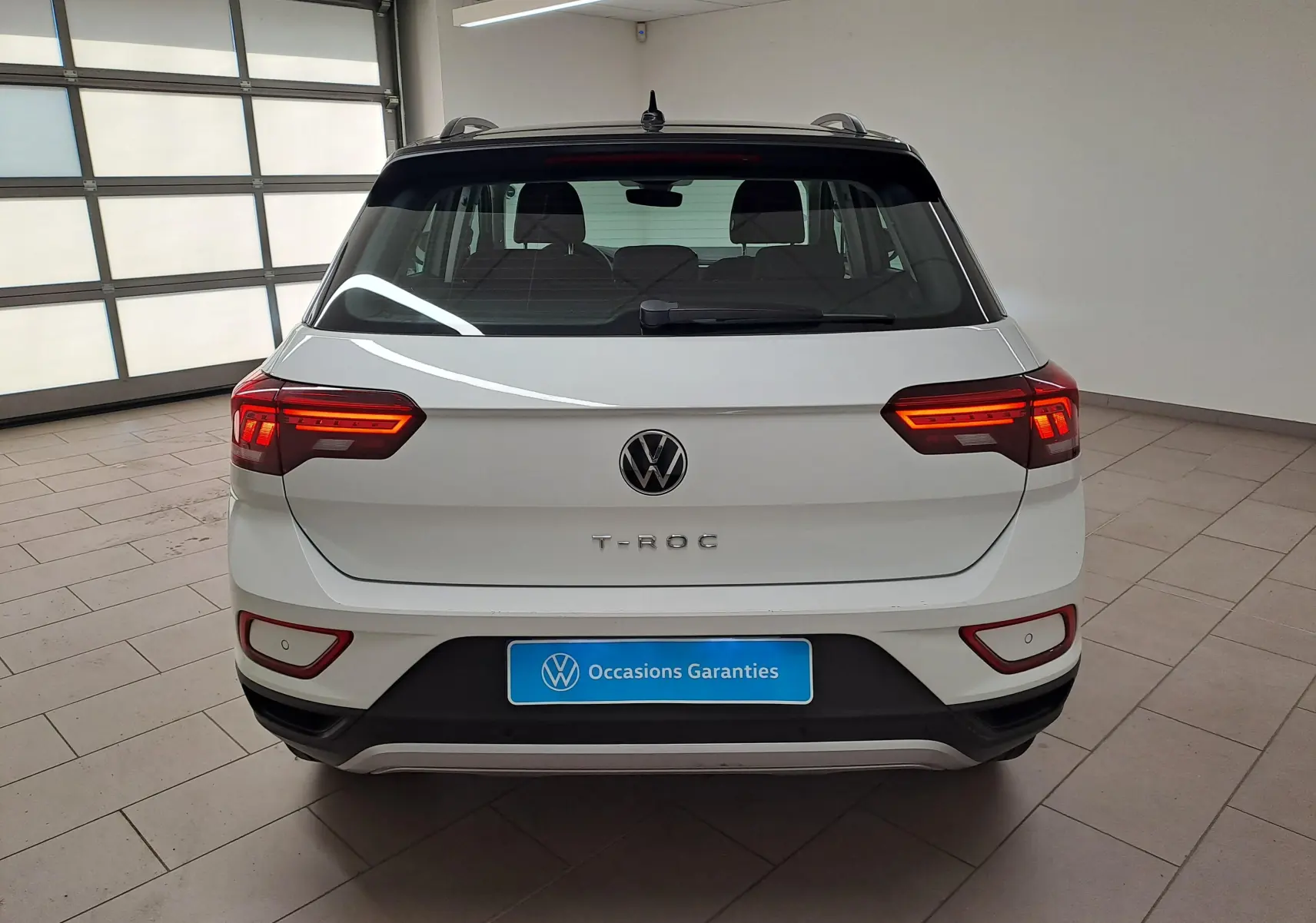 Vue arrière d'un Volkswagen T-Roc blanc pur avec toit noir, feux allumés, dans un garage lumineux.