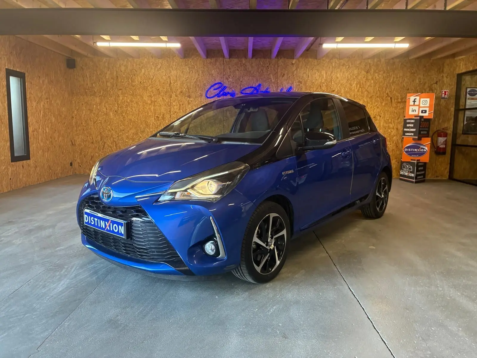 Toyota Yaris hybride bleu en 3/4 avant droit, avec jantes noires et toit noir dans un garage intérieur.