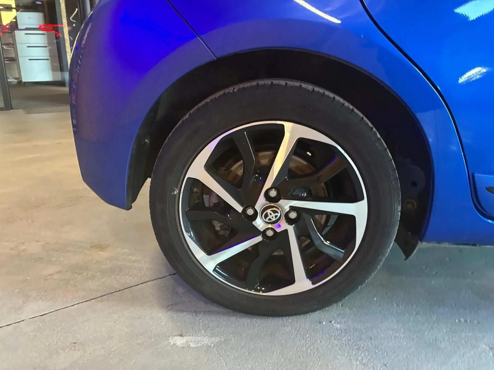Gros plan sur la roue arrière droite d'une Toyota Yaris bleu vif avec jante alliage bicolore en intérieur d'atelier.