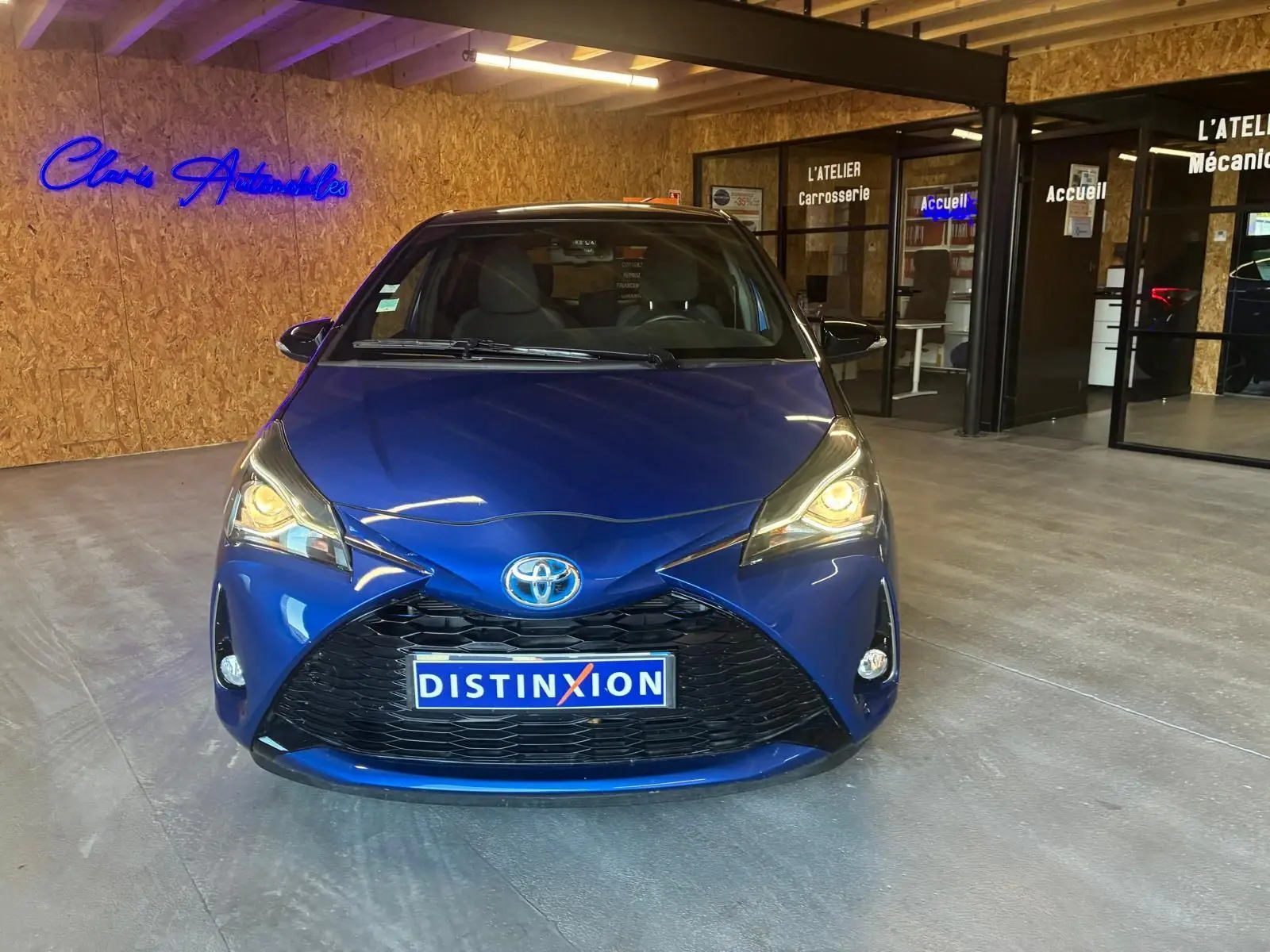 Toyota Yaris hybride bleu vue de face en intérieur, phares allumés et plaque Distinxion visible.