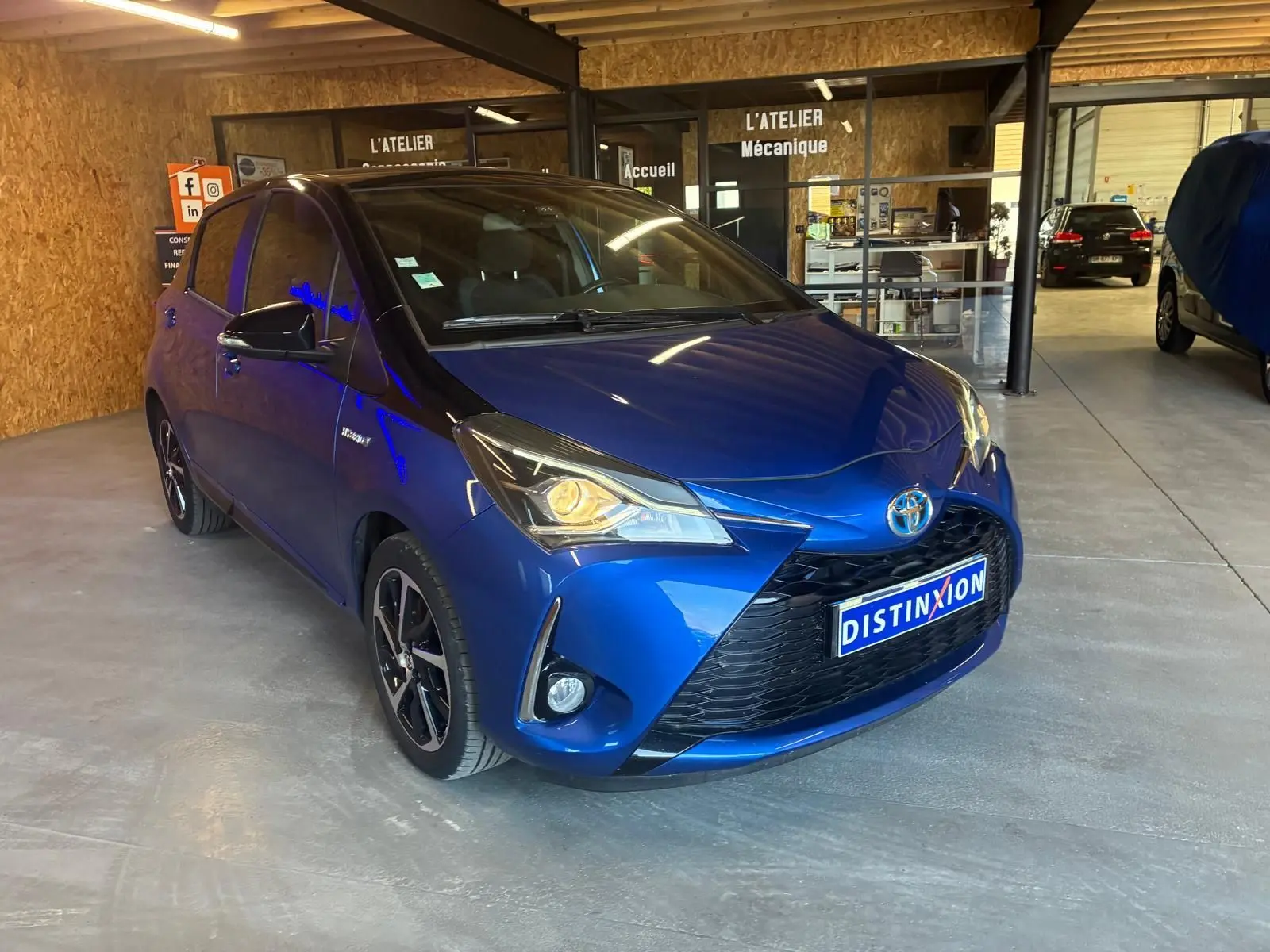 Toyota Yaris hybride bleu vue 3/4 avant droit dans un garage avec phares allumés et jantes noires.