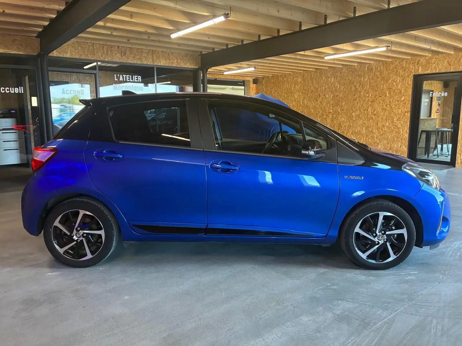 Profil côté gauche d'une Toyota Yaris hybride bleue 5 portes, avec jantes noires et toit noir en intérieur d'atelier.
