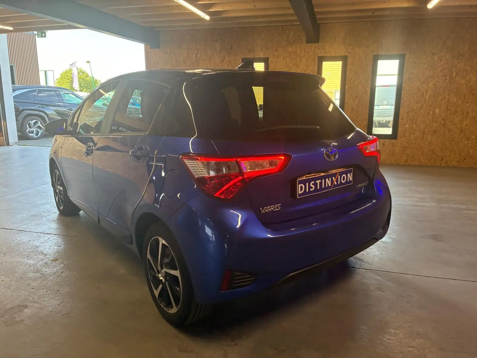 Toyota Yaris hybride bleu vue 3/4 arrière droit dans un garage, avec feux arrière allumés et jantes noires.