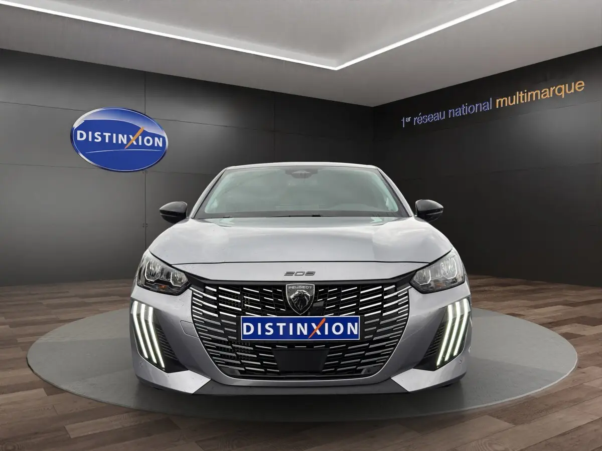 Vue frontale d'une Peugeot 208 gris clair avec calandre noire et feux LED verticaux allumés en showroom.