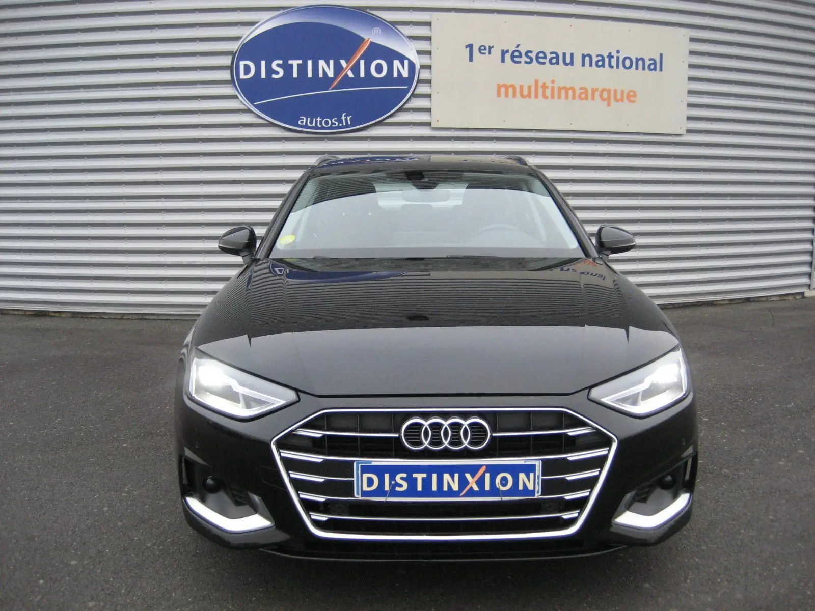 Vue frontale d'une Audi A4 Avant noire 2022 avec phares allumés et calandre chromée, devant un mur métallique.