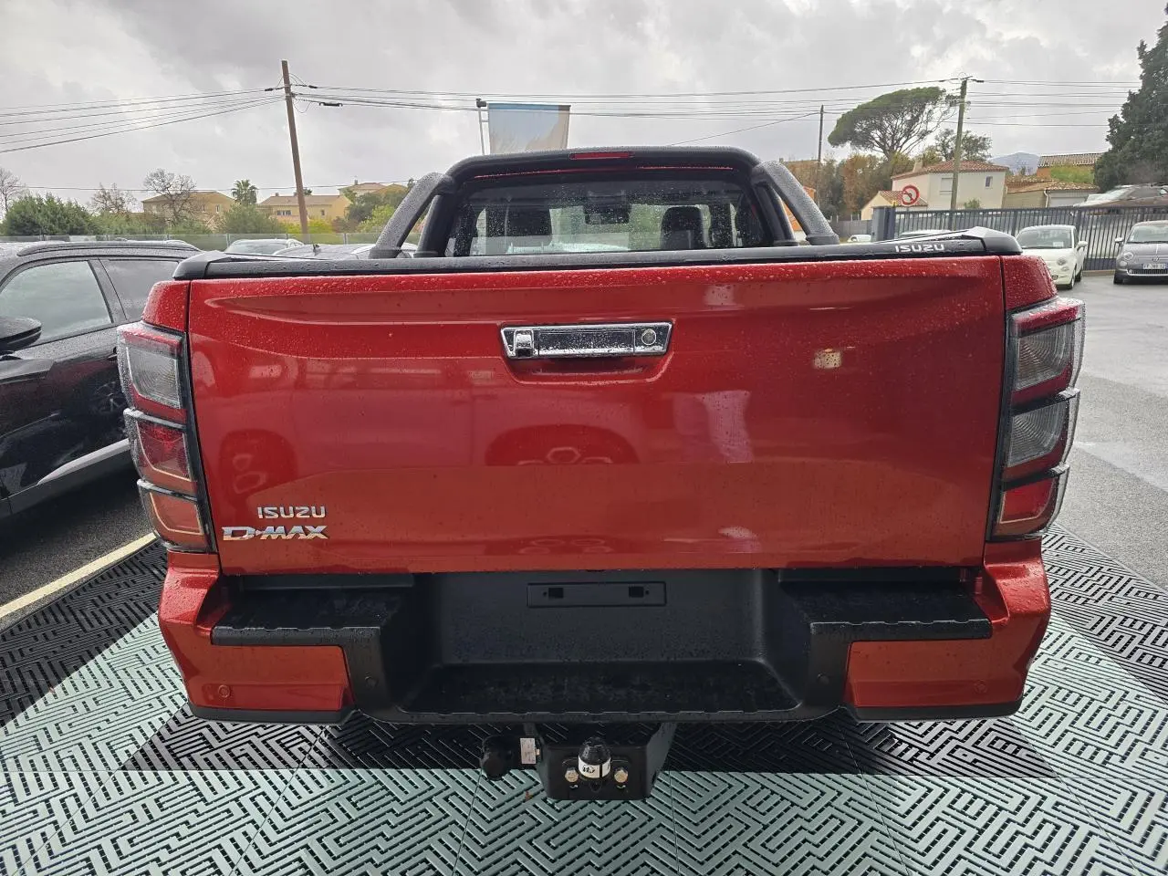 Vue arrière d'un pick-up Isuzu D-MAX orange métallisé avec attelage et hayon fermé sous la pluie.