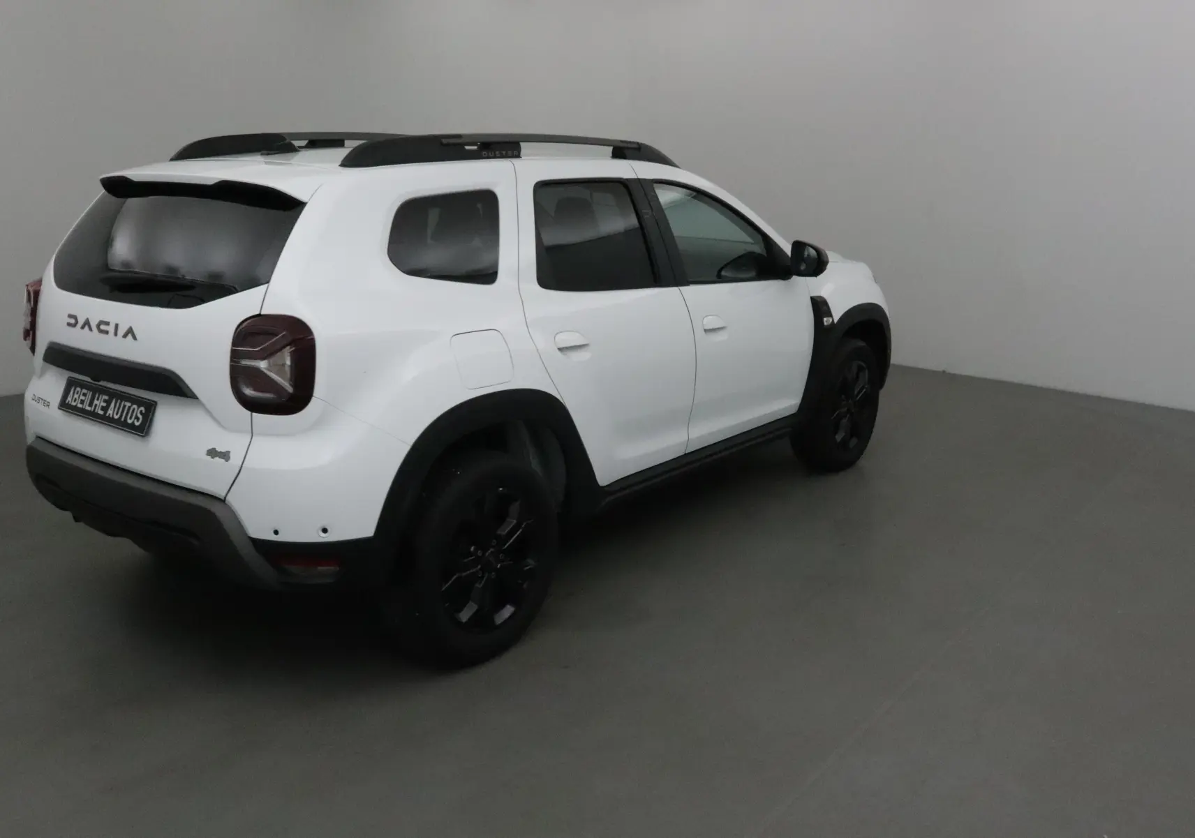 Dacia Duster 4x4 blanc en 3/4 arrière droit, avec jantes noires et barres de toit visibles dans un intérieur neutre.