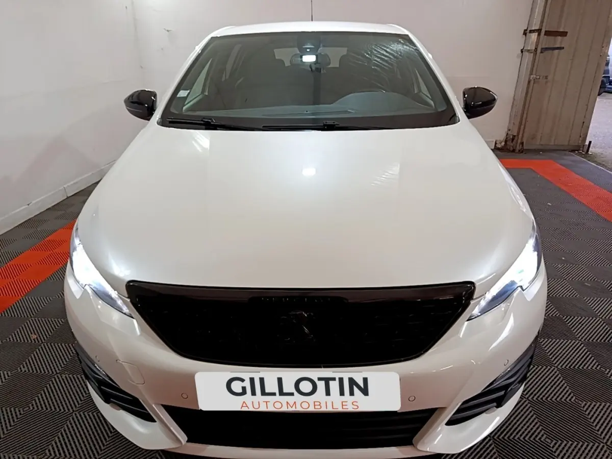 Vue avant d'une Peugeot 308 blanche avec calandre noire et phares LED allumés dans un garage.