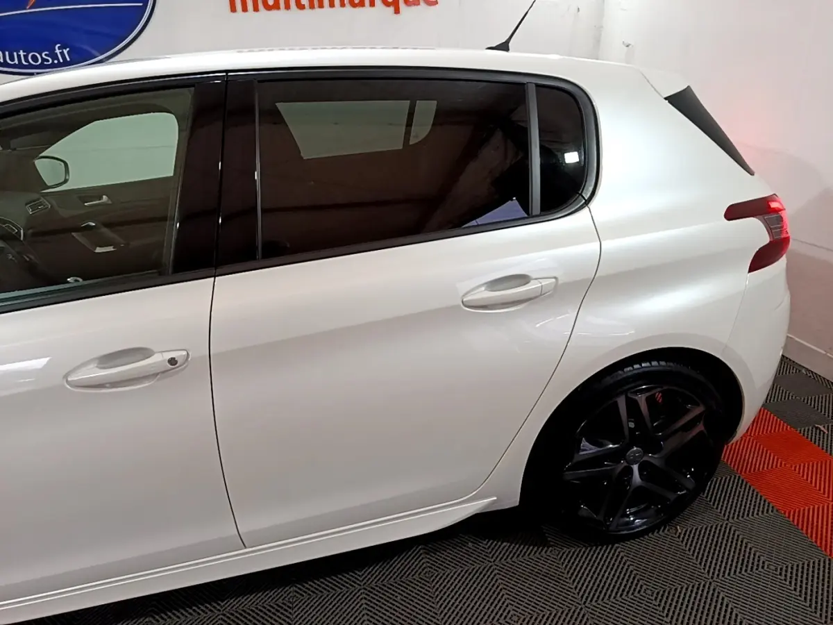 Profil côté gauche d'une Peugeot 308 blanche avec jantes noires et vitres teintées en intérieur showroom.