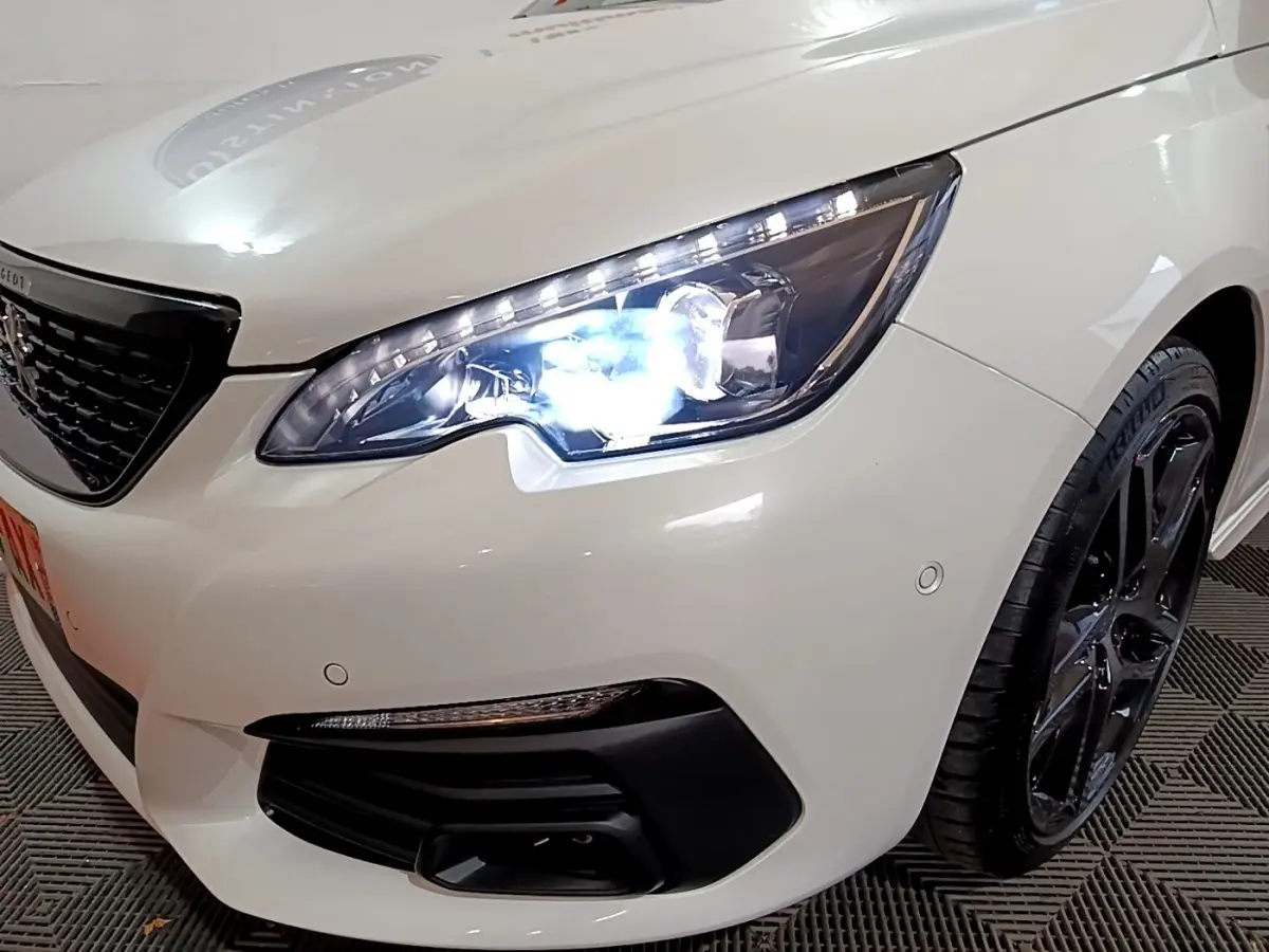 Gros plan sur l'avant droit blanc d'une Peugeot 308 2019 avec phare LED allumé et jante noire brillante